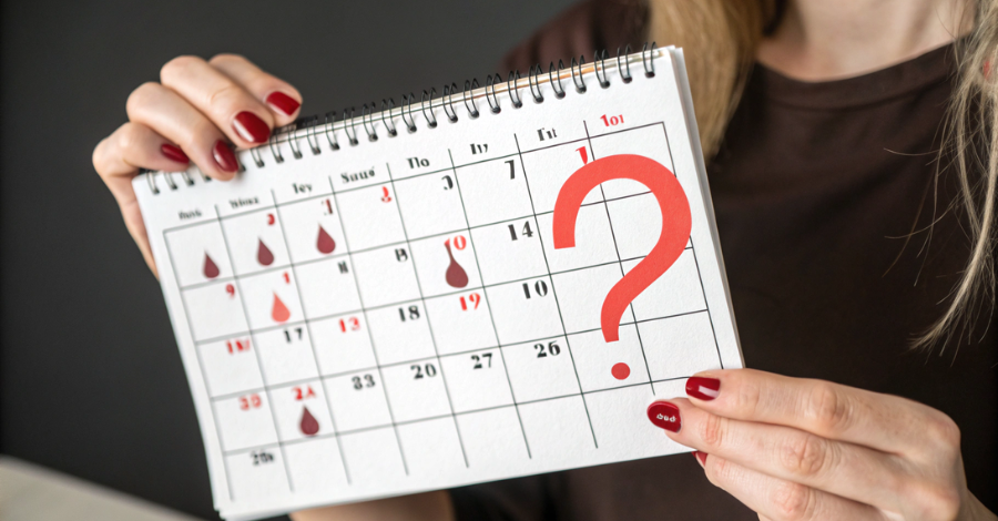 Mão de mulher em frente a um calendário menstrual, com um ponto de interrogação sobre a data, representando um sinal de alerta de saúde.